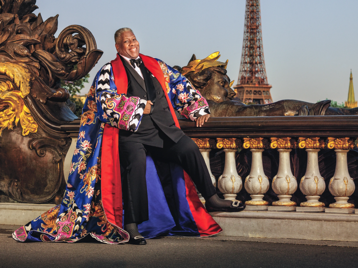 Photo of Andre Leon Talley in a black tuxedo with a red, blue, and multi-colored, floor-length robe over the suit. He is outside with what appears to be the Eiffel Tower in the distance behind him.