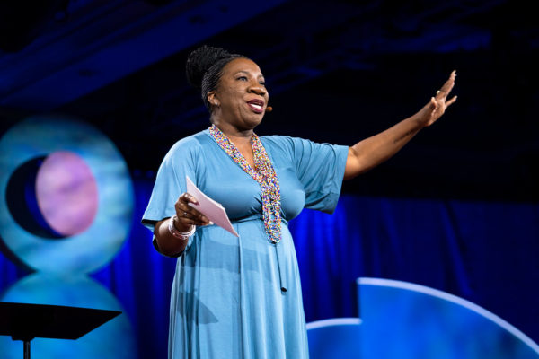 Tarana Burke giving a TED talk, wearing a blue dress, with her arms outstretched